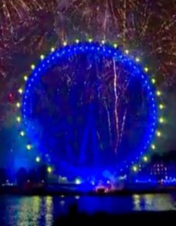 The London Eye, a ferris wheel, lit up in blue with yellow lights round the outside (as on the European flag). Traces of other fireworks can be seen around it. The lights are reflected in the River Thames at the bottom.