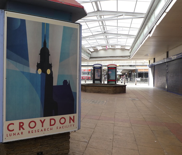 A picture of a 1970s poster showing a clock tower with lights shining from two sides, with the words in red underneath, "Croydon lunar research facility", in a covered walkway with shuttered shops on the right-hand side and two BT phone boxes in the background.