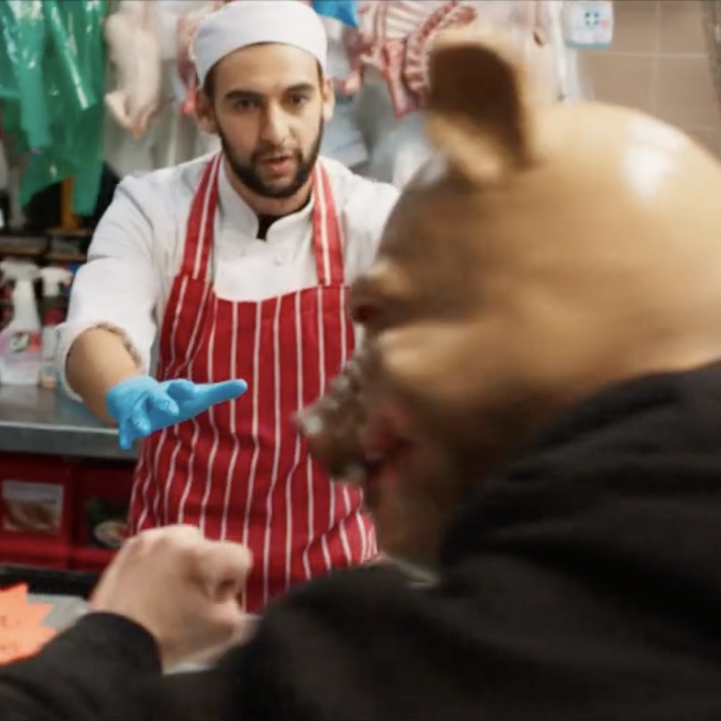 A South Asian man wearing white butcher's clothes, a red and white striped apron and blue plastic gloves tries to calm a man wearing a pig mask who is threatening him. An electronic scale and hanging cuts of meat are behind him.
