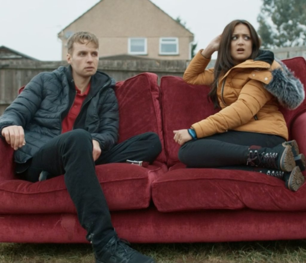 Gethin, a young white man wearing a black jacket and trousers and a red T-shirt sits on a red sofa in the street with a young South Asian woman who is wearing jeans and a yellow jacket. There is a concrete wall behind them, and a new red-brick house behind that.