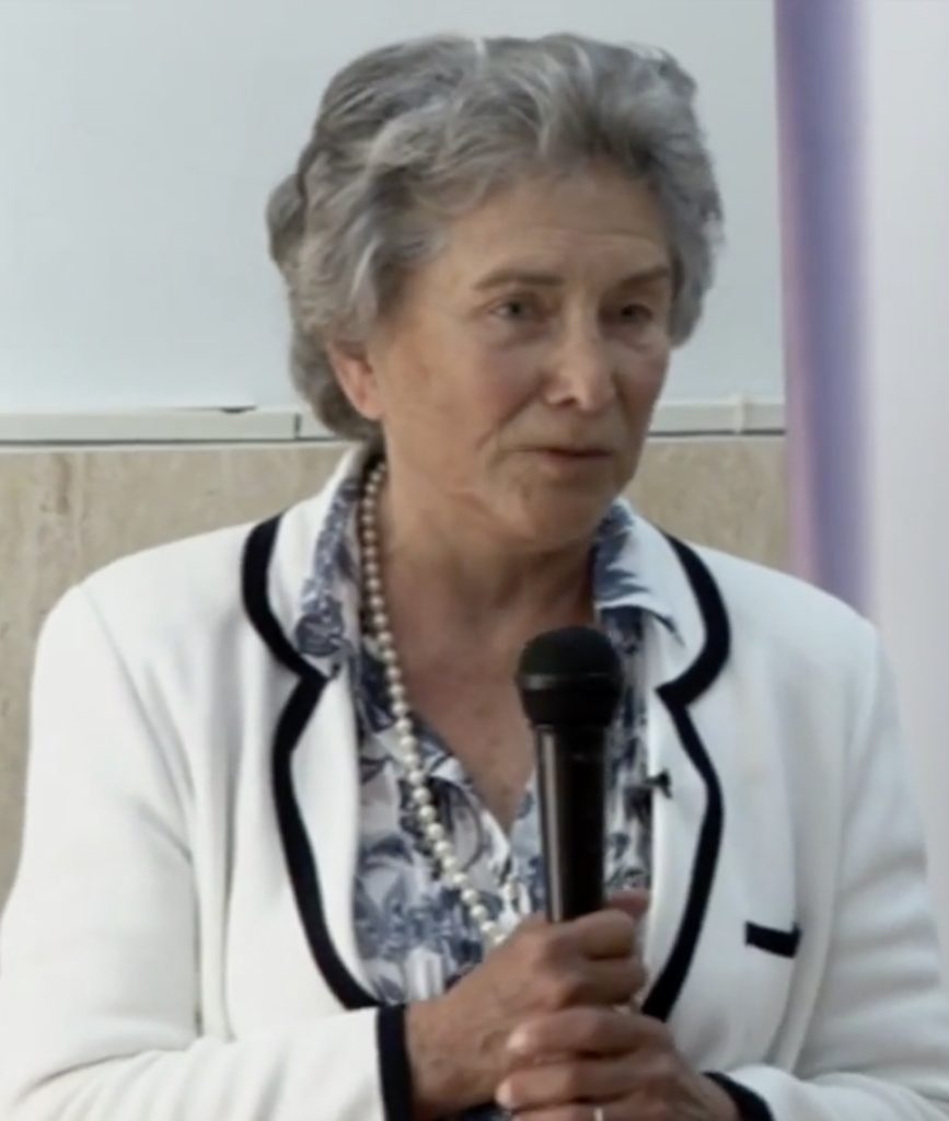 Picture of an elderly white woman with grey hair wearing a blue floral blouse under a white jacket with black edging, holding a black microphone in her hand.