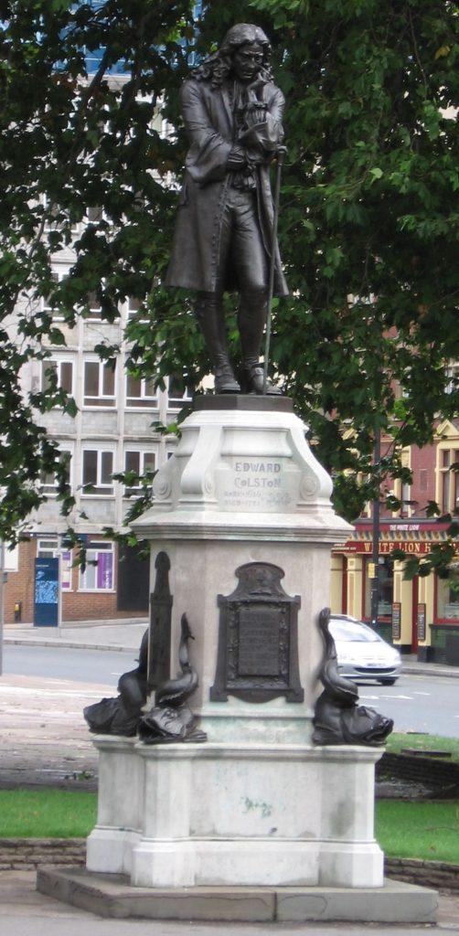 A bronze statue of Edward Colston atop a stone monument with four figures on a lower plinth.