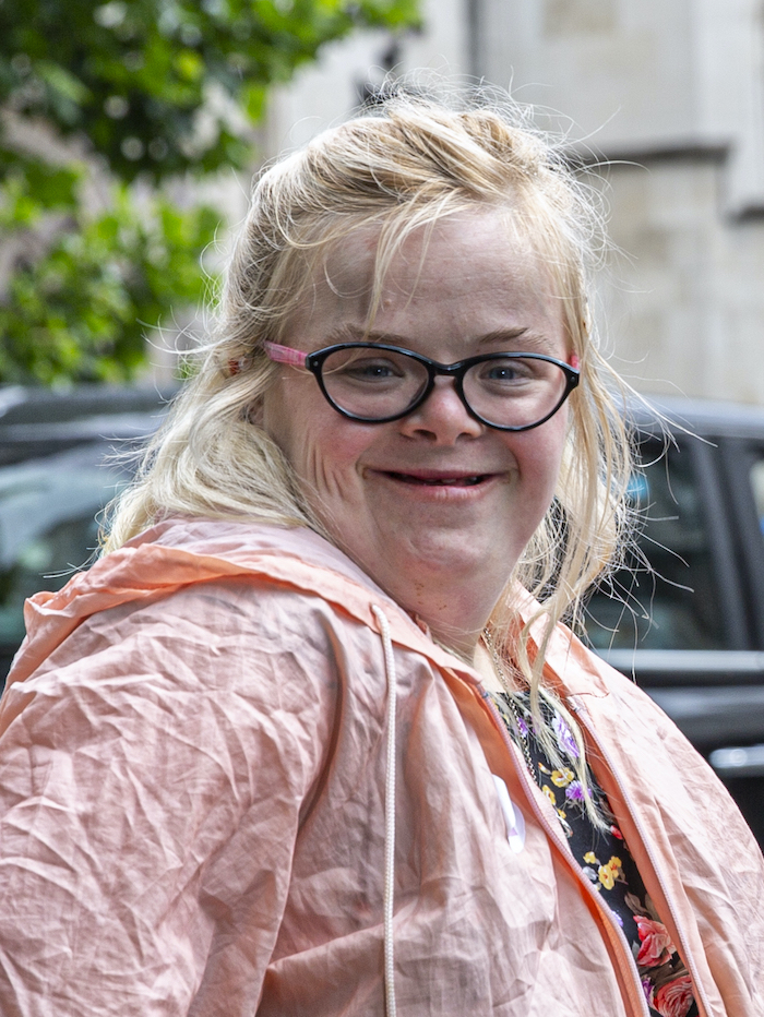 Picture of Heidi Crowter, a white woman with Down's syndrome wearing glasses with thick black rims and a pink raincoat and a black top with different coloured flowers on underneath it. A car, a tree and a stone building are behind her, but are blurred.