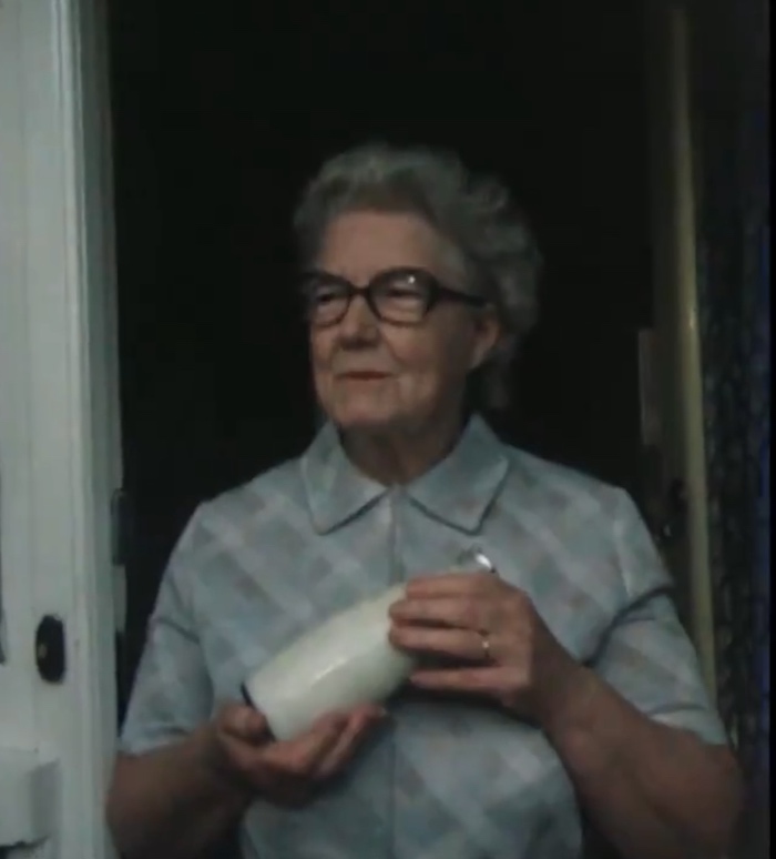 An old white lady standing at her front door, holding a one-pint glass milk bottle.
