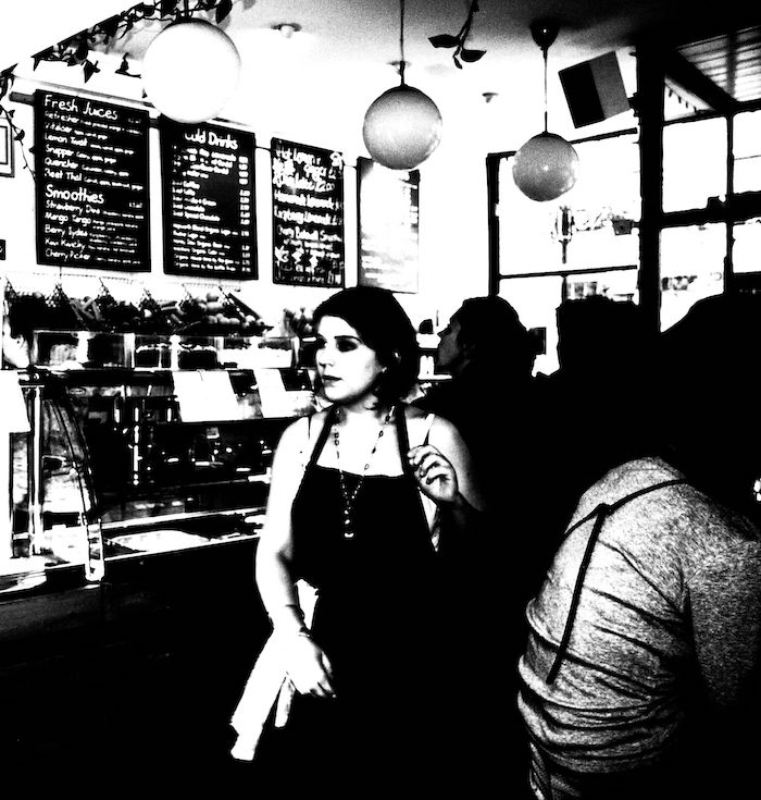 A black-and-white photograph of a cafe with a handwritten menu on the back wall and coffee machines below it. Customers are sitting around on the right and a young female member of staff is seen facing to the left.