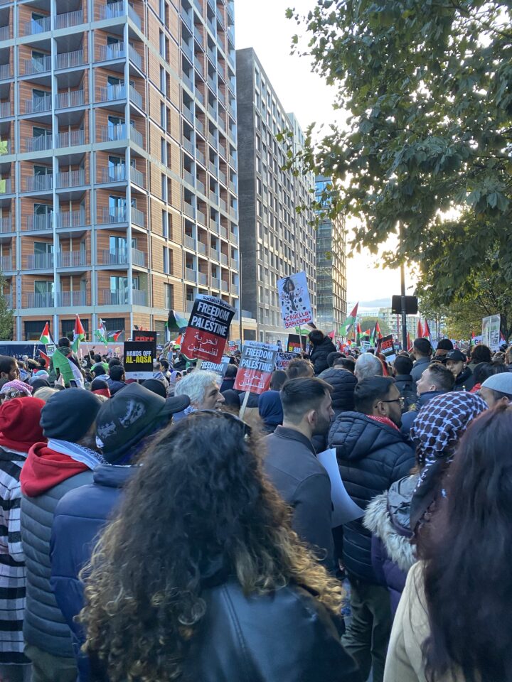 A demonstration in London, with people holding banners with "Free Palestine" type slogans. Some new blocks of flats or offices are visible in the background.
