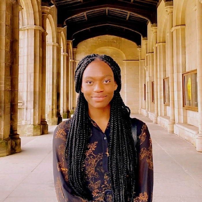 Picture of Stella Maris, a Black woman in her 20s with long, braided black hair wearing a purple and gold blouse, standing in a wide stone passageway with arches to the left and portraits on the stone walls to the right.