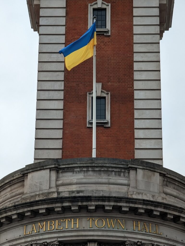 A red-brick tower rising from a platform at the top of a building. On the platform is a flagpole with a Ukrainian blue and yellow flag flying from it. Below the platform are the words "Lambeth Town Hall" in golden Roman lettering on the grey stone.
