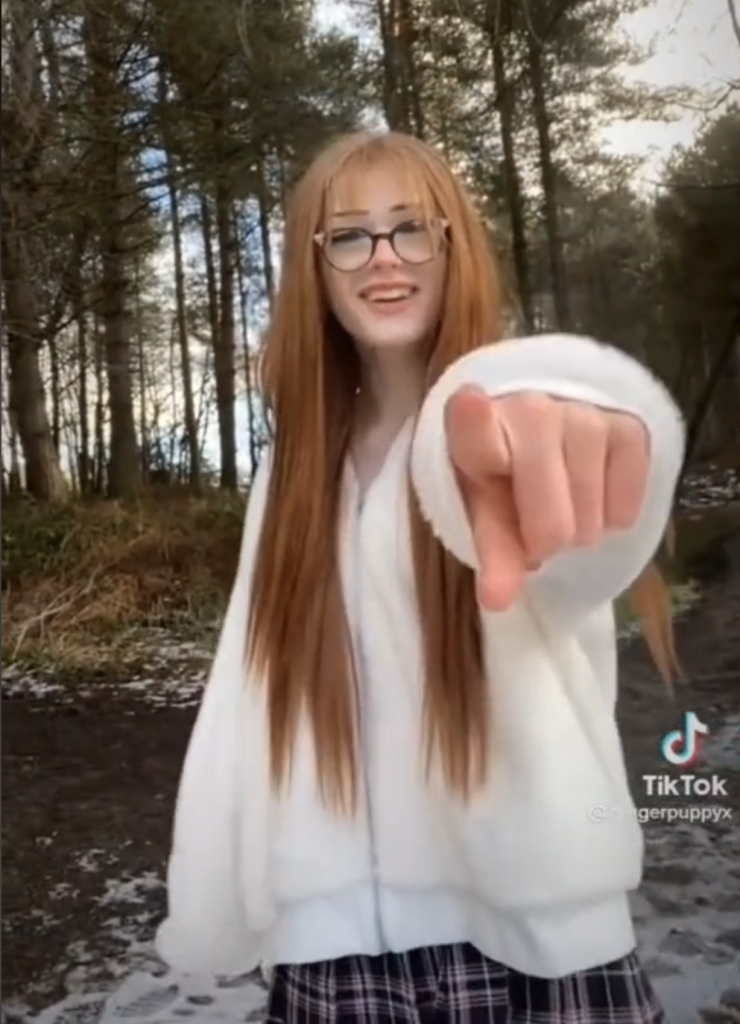 Picture of Brianna Ghey, a young, white, female presenting teenager with long, blonde hair and glasses, standing in a wooded area wearing a white cardigan and tartan school skirt with her finger pointed out towards the camera.