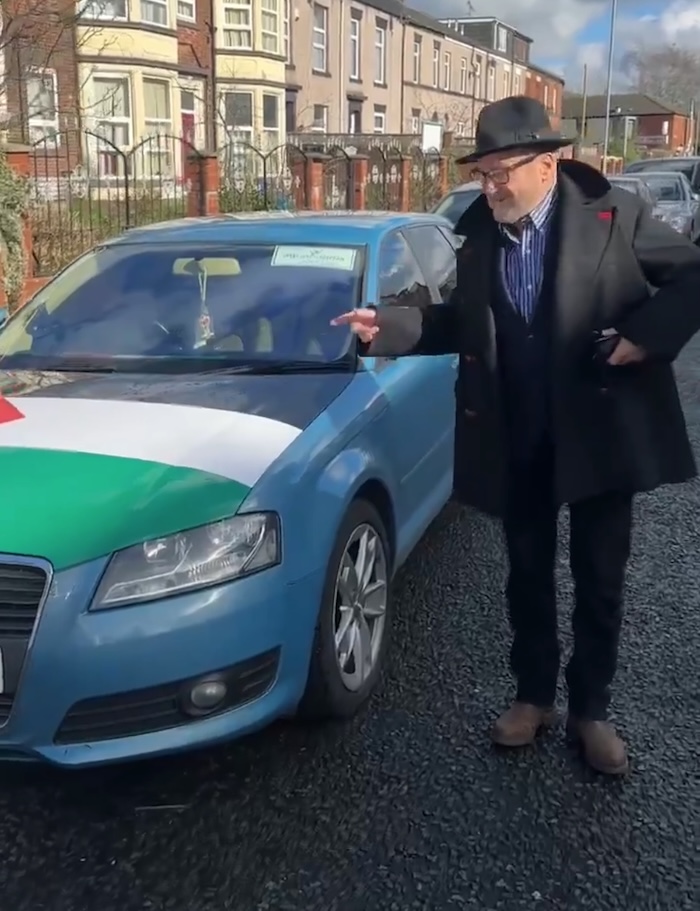 George Galloway, an elderly white man with a short white beard, wearing a black rimmed hat, a black blazer and trousers with a blue and white striped shirt underneath, admires a car (a blue Audi) with a Palestinian flag printed on its bonnet.