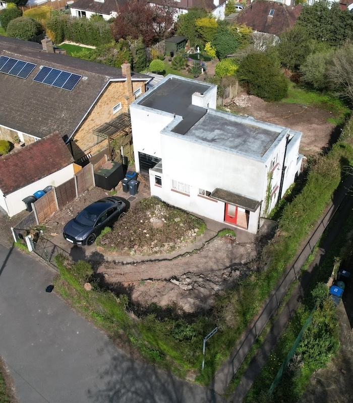 An aerial photo of a small white house with a flat roof. There are small windows on the ground floor and none on the storey above. The front garden has a black saloon car in it but its front and rear gardens are in a poor condition.