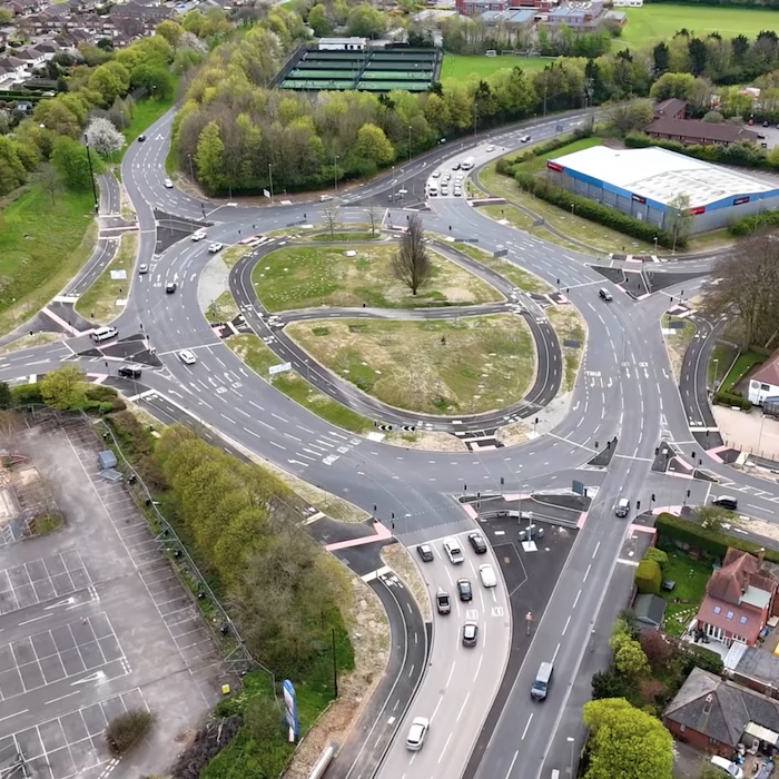 An aerial view of a large traffic roundabout with six roads meeting. Around it are fields, houses, a large big-box store and a big car park. Inside the roundabout is a cycle track running just inside the main roadway, and another runs across it.