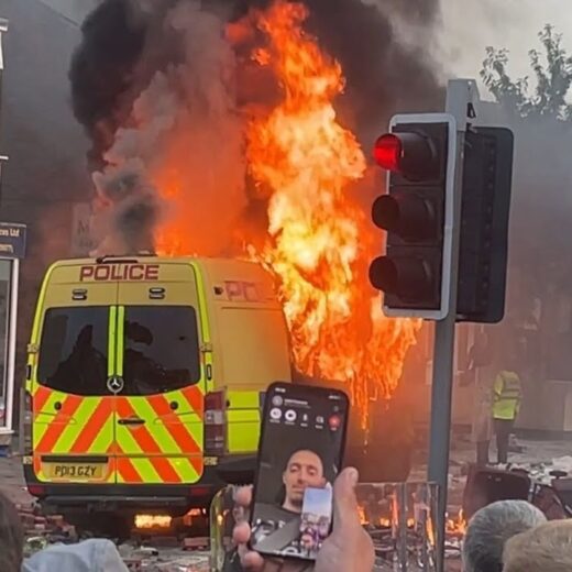 A yellow police van on fire in a street. In the foreground, a man relays the scene to his friend through his mobile phone.