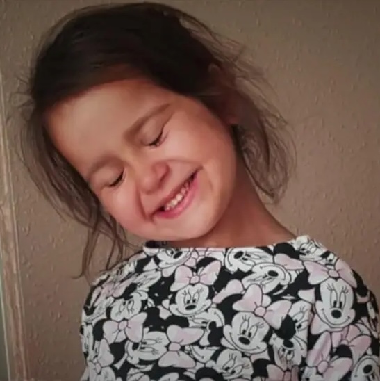 Picture of Sara Sharif, a young, white appearing girl with dark brown hair, wearing a top with a cartoon pattern. Her head is tilted to one side, her eyes are closed and she is smiling.