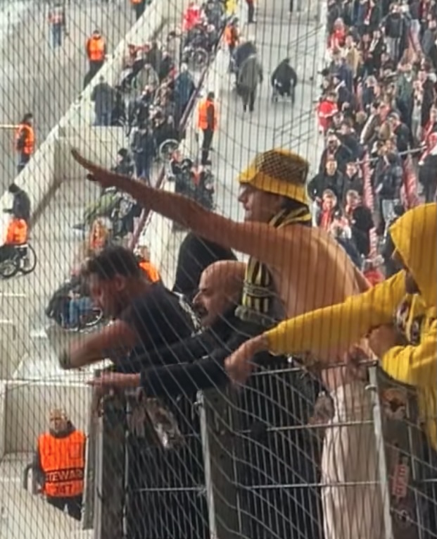 A man with a yellow and blue hat and no top on gives a Nazi salute from behind a crowd-control fence in a football stadium.