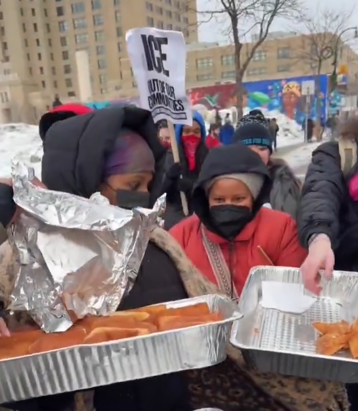 A street in Minneapolis where Somali women with metal trays of samosas are giving them out to protesters. An anti-ICE banner can be seen in the background.