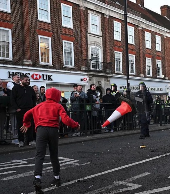 A photo of a demonstration in an English street. A boy in a red hoodie has thrown a red traffic cone at riot police who are facing him with clear plastic shields in front of them. Men stand on the pavement watching. Behind them is a long red-brick façade; one part of the building houses the HSBC bank and another the Waterstone's bookshop.