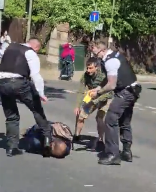 Two police officers and a third man in a khaki jacket stand over a man who is on the floor. One of the officers is kicking the man in the head and the other is holding a yellow Taser stun-gun.