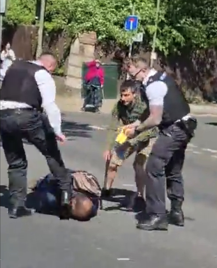 Two police officers and a third man in a khaki jacket stand over a man who is on the floor. One of the officers is kicking the man in the head and the other is holding a yellow Taser stun-gun.