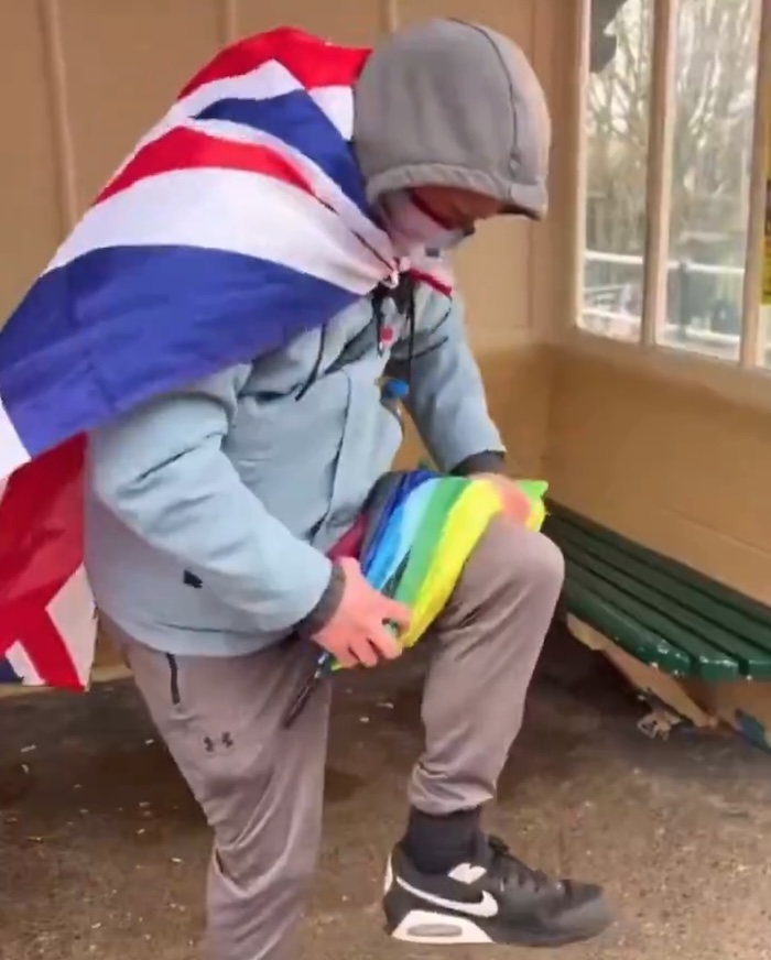A man wearing a Union Jack flag draped over his back, standing in a beach shelter, breaks a rainbow umbrella over his knee.