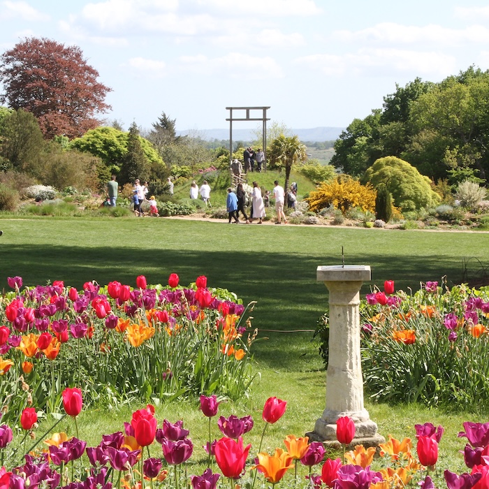 A lawn in a large country garden. In the foreground is a sundial surrounded by red, purple and yellow tulips. In the background is a rock garden, with various small trees and a wooden viewing platform. Behind that is a view into the Sussex countryside.
