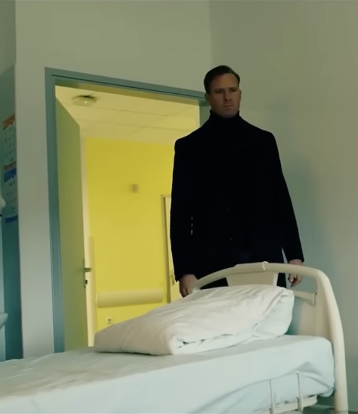 A still showing Armie Hammer walking into a hospital room wearing a dark jacket. The door is open behind him and a hospital corridor can be seen; in front of him is an empty hospital bed.