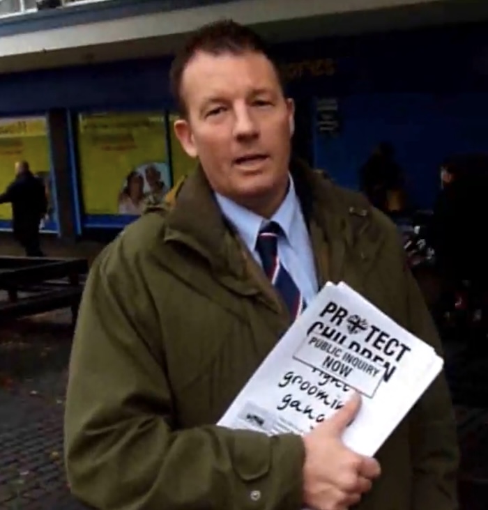 A man in a suit and tie with a green jacket holds a stack of pieces of papers saying "Fight Grooming Gangs, Protect Children, Public inquiry now!".