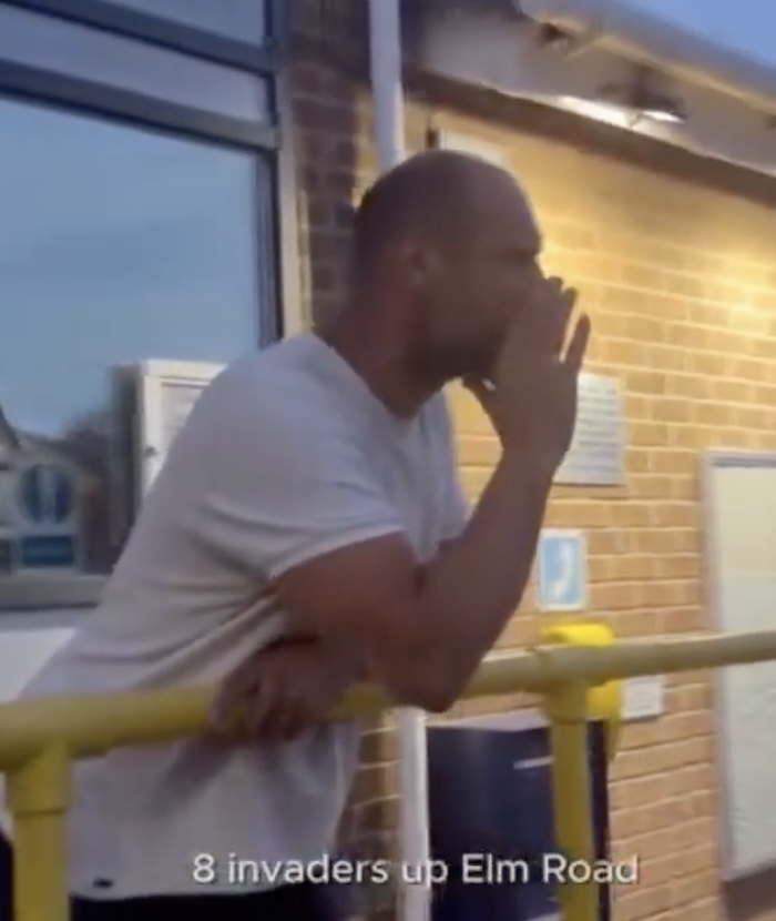 A man standing against a yellow railing outside a building, telling a crowd "there are eight invaders up Elm Road".