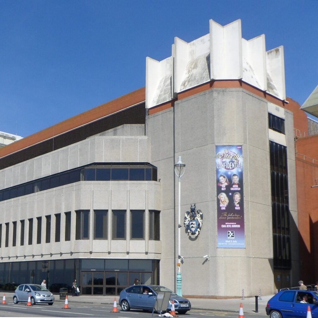 Picture of the Brighton Centre, a conference and exhibition centre in Brighton. It is a concrete building with a glass ground-floor facade and a line of vertical first-floor windows. A coat of arms is visible as is a poster for a 2013 event, Strictly Confidential (related to Strictly Come Dancing, a UK TV ballroom dancing competition).